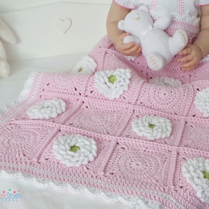May include: A pink crocheted baby blanket with white flowers and a white crocheted border. The blanket is on a white surface.