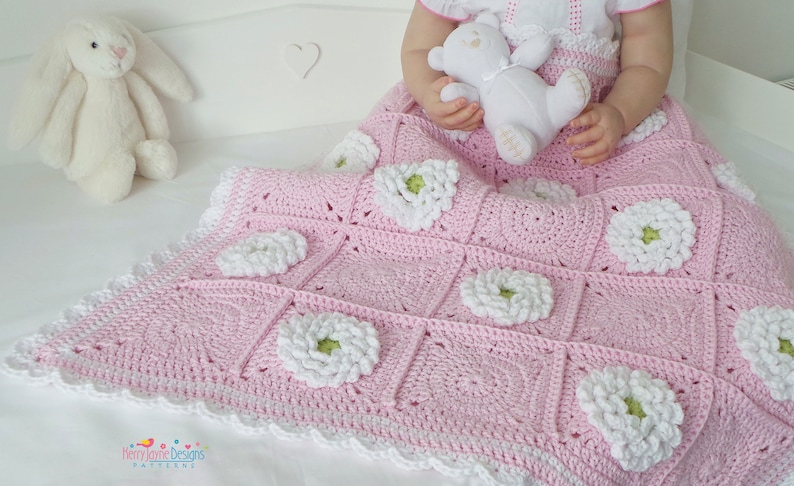 May include: A pink crocheted baby blanket with white flowers and a white crocheted border. The blanket is on a white surface.
