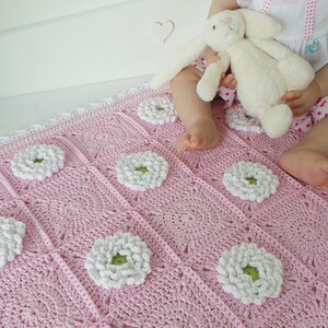 May include: A pink crocheted baby blanket with white daisy flowers. The blanket has a white crocheted border.