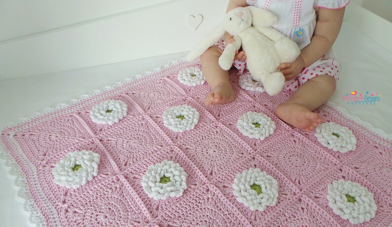 May include: A pink crocheted baby blanket with white daisy flowers. The blanket has a white crocheted border.
