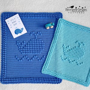 Puede incluir: Dos mantas de bebé de ganchillo en tonos azules, con un diseño de ballena. Una manta es azul oscuro, la otra es azul claro. Una tarjeta que dice "It's a Baby Boy" con una ilustración de ballena está en la manta azul oscuro.