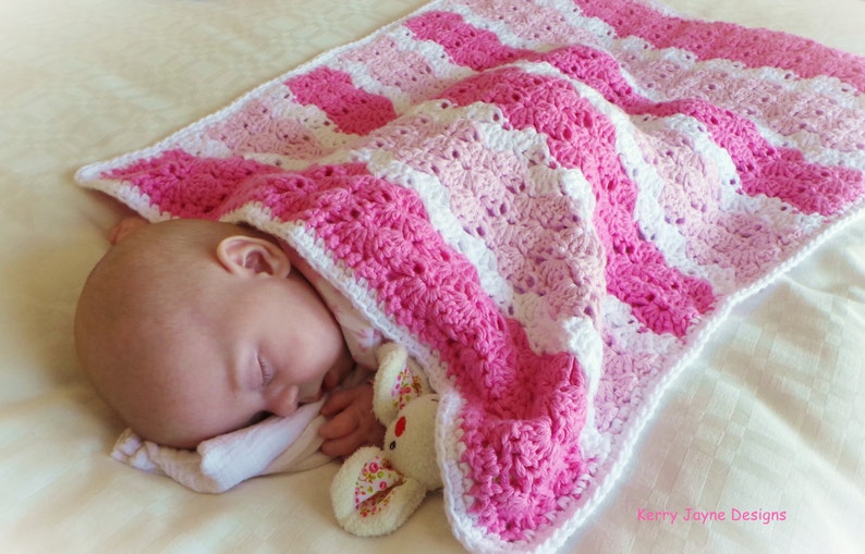 Peut inclure: Une couverture de b&eacute;b&eacute; en crochet ray&eacute;e rose et blanche avec un b&eacute;b&eacute; endormi dessus. Le b&eacute;b&eacute; tient un petit animal en peluche.