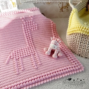 May include: A pink crocheted baby blanket with a giraffe design. The blanket has a textured border and a small stuffed giraffe toy is sitting on top of it.