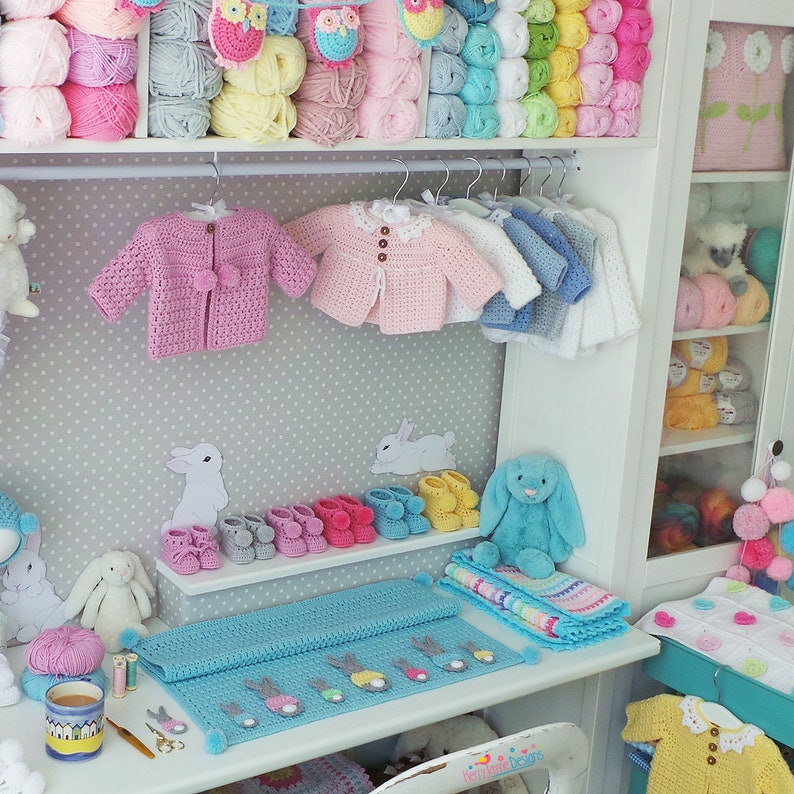 May include: A white shelf unit with a variety of crocheted items, including baby clothes, blankets, and toys. The shelf is decorated with a gray and white polka dot wallpaper. There are also several balls of yarn in various colors on the shelf.