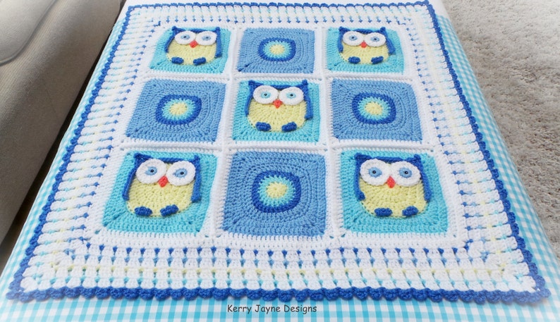 May include: A crocheted baby blanket with a blue, white, and yellow color scheme. The blanket features a repeating pattern of owls with large eyes. The blanket is bordered with a white and blue trim.