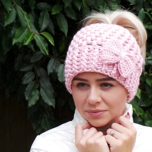 May include: A pink crocheted hat with a ponytail hole and a bow on the side.