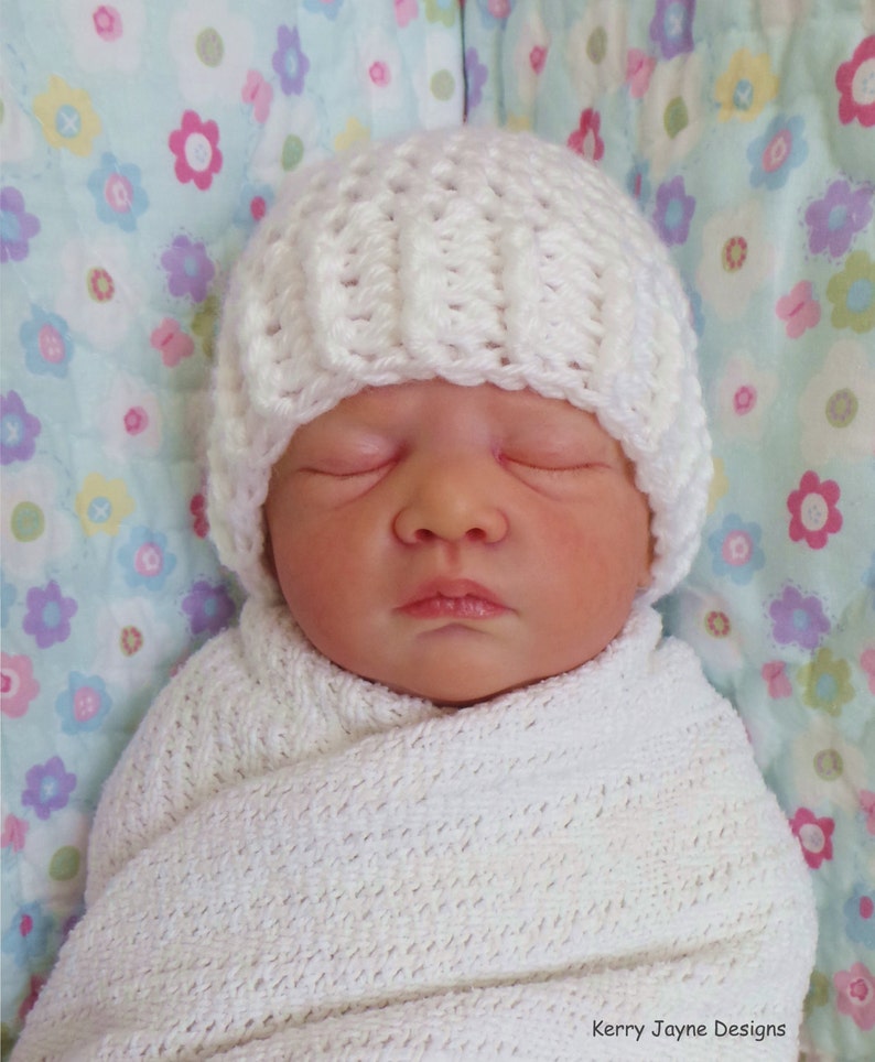 May include: A white crocheted baby hat with a simple stitch pattern. The hat is on a baby wrapped in a white blanket with a floral pattern.