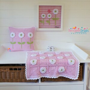 May include: A pink and white crocheted baby blanket with a flower design. The blanket is on a white changing table with a wicker basket underneath. There is a pink and white pillow with a flower design on the changing table. A framed picture with a pink and white gingham background and white flowers is on the wall above the changing table. The text "Kerry Jayne Designs PATTERNS" is on the wall.