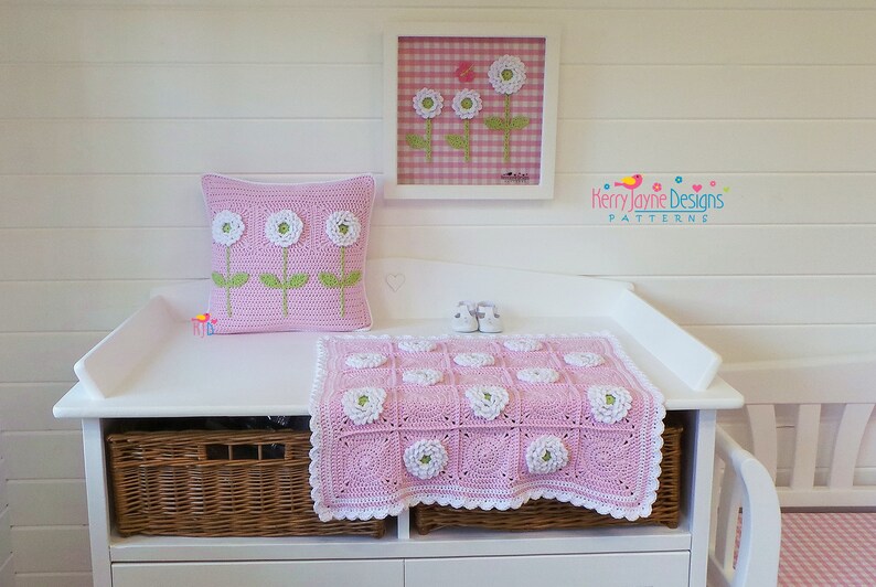 May include: A pink and white crocheted baby blanket with a flower design. The blanket is on a white changing table with a wicker basket underneath. There is a pink and white pillow with a flower design on the changing table. A framed picture with a pink and white gingham background and white flowers is on the wall above the changing table. The text "Kerry Jayne Designs PATTERNS" is on the wall.