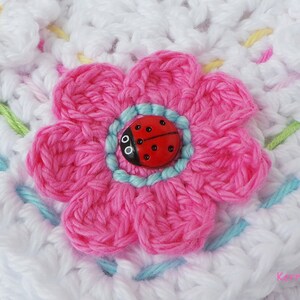 May include: A close-up of a crocheted flower with a pink center and a ladybug button in the middle. The flower is surrounded by white crocheted fabric with a light blue border.