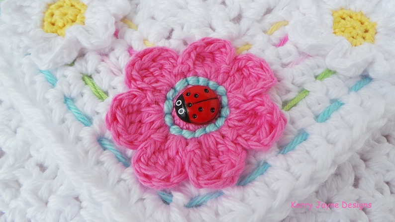 May include: A close-up of a crocheted flower with a pink center and a ladybug button in the middle. The flower is surrounded by white crocheted fabric with a light blue border.