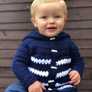May include: A toddler wearing a blue knitted cardigan with white stripes and white buttons. The cardigan has a hood.