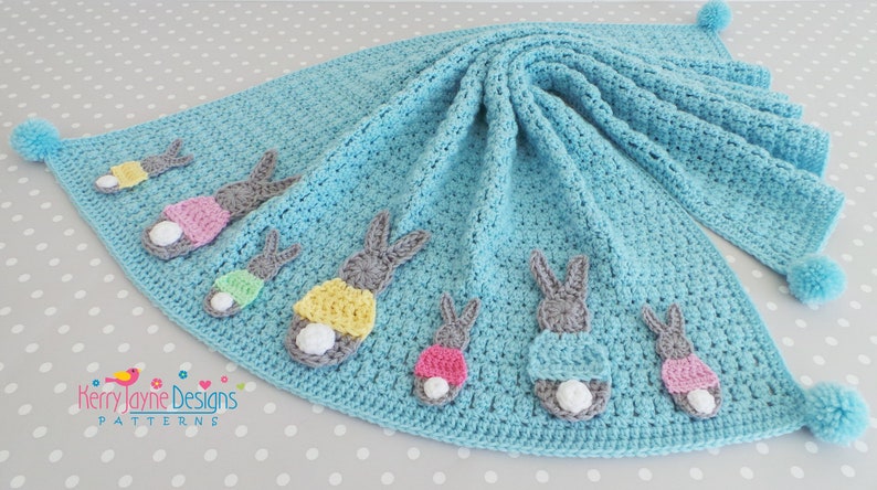 May include: A light blue crocheted baby blanket with a scalloped edge and six bunny appliques in various colors. The blanket has pom-poms on the corners.