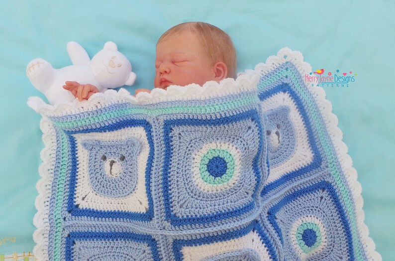 K&ouml;nnte beinhalten: Eine blaue und wei&szlig;e geh&auml;kelte Babydecke mit einem B&auml;renmuster. Die Decke hat einen wei&szlig;en geh&auml;kelten Rand mit einem gewellten Saum. Ein wei&szlig;er Teddyb&auml;r liegt auf der Decke neben einem Baby.
