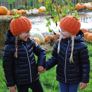 May include: Two children wearing orange pumpkin hats with green stems, smiling and holding hands in a field of pumpkins.