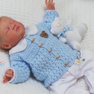 May include: A light blue crocheted baby cardigan with white trim, brown buttons, and a brown felt bear applique. The cardigan is worn by a baby doll with blonde hair.