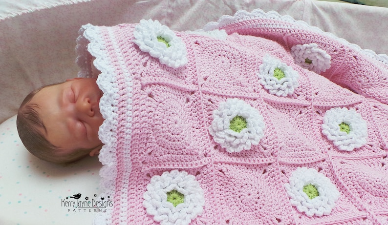 May include: A pink crocheted baby blanket with white and green flowers. The blanket is made with a granny square pattern and has a white crocheted border.