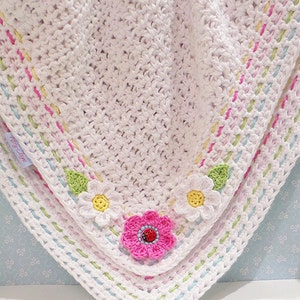 May include: White crocheted baby blanket with a colorful border and crocheted flower accents. The flowers are pink, white, and yellow with green leaves. The blanket has a label that says "Pretty Little"