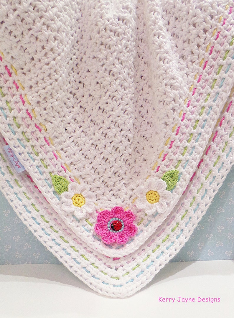 May include: White crocheted baby blanket with a colorful border and crocheted flower accents. The flowers are pink, white, and yellow with green leaves. The blanket has a label that says "Pretty Little"