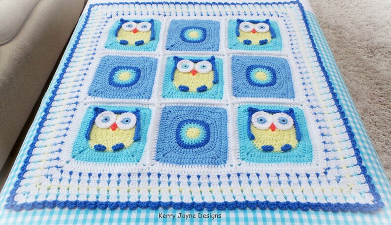 May include: A crocheted baby blanket with a blue, white, and yellow color scheme. The blanket features a repeating pattern of owls and circles. The blanket is bordered with a blue and white trim.