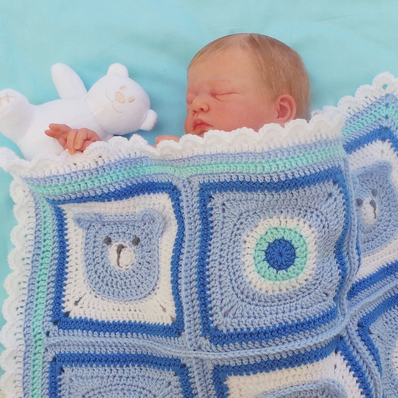 K&ouml;nnte beinhalten: Ein Baby schl&auml;ft auf einer blauen und wei&szlig;en geh&auml;kelten Babydecke mit einem wei&szlig;en Teddyb&auml;ren. Die Decke hat einen gewellten Rand und zeigt einen blauen B&auml;ren und ein blaues Kreisdesign.