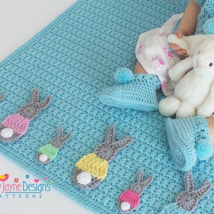 May include: A blue crocheted baby blanket with a row of five gray bunny appliques. Each bunny has a different colored tummy. The blanket is on the floor and a baby is sitting on it. The baby is wearing a white dress with pink flowers and a blue crocheted cardigan with pom poms. The baby is holding a white stuffed bunny.