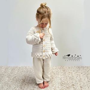 May include: A cream-coloured long-sleeve crochet cardigan featuring a textured bobble stitch pattern along the cuffs and hem, fastened with five light brown wooden buttons, paired with matching cream-coloured lightweight cotton trousers.