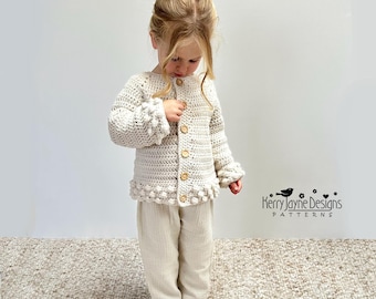 May include: A cream-colored long-sleeve crochet cardigan featuring a textured bobble stitch pattern along the cuffs and hem, fastened with five light brown wooden buttons, paired with matching cream-colored lightweight cotton trousers.