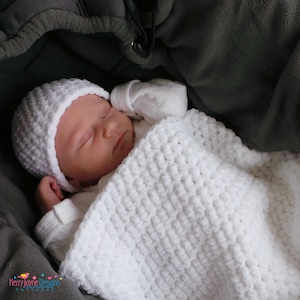 May include: A white crocheted baby blanket with a white crocheted hat on top. The baby is sleeping peacefully under the blanket.
