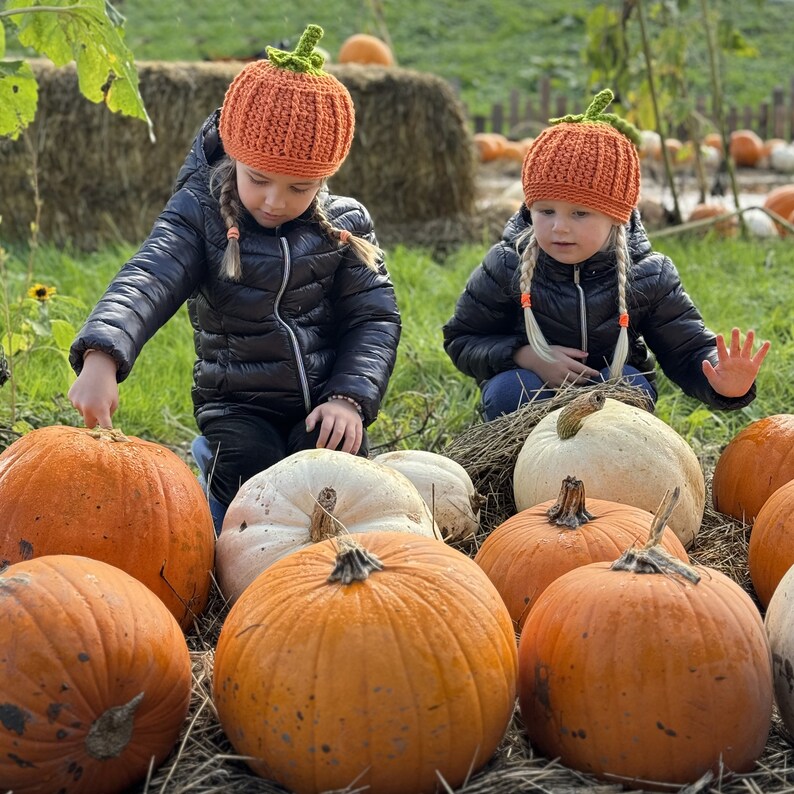 May include: Two children wearing orange and brown knitted hats are standing in a field of pumpkins. The pumpkins are various sizes and colors, including orange, white, and green.
