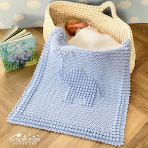 Peut inclure: Une couverture pour bébé en crochet bleu clair avec un motif d'éléphant texturé. La couverture a une bordure blanche avec de petits pompons autour des bords.