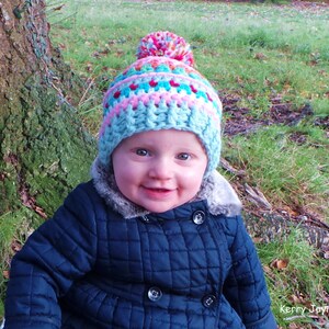 May include: A young child wearing a blue quilted jacket and a colorful crocheted hat with a pom-pom on top. The hat has stripes of pink, blue, and white.