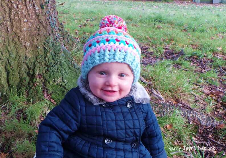 May include: A young child wearing a blue quilted jacket and a colorful crocheted hat with a pom-pom on top. The hat has stripes of pink, blue, and white.
