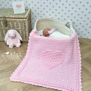 Puede incluir: Una manta de bebé de crochet rosa con un diseño en forma de corazón. La manta tiene un borde blanco y está hecha con un punto texturizado. La manta está sobre un suelo de madera.
