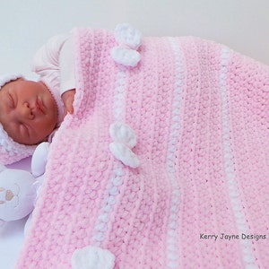 K&ouml;nnte beinhalten: Eine gestrickte Babydecke in rosa und wei&szlig; mit wei&szlig;en geh&auml;kelten Blumen. Die Decke ist &uuml;ber einem Baby gefaltet, das eine rosa und wei&szlig;e geh&auml;kelte M&uuml;tze tr&auml;gt.