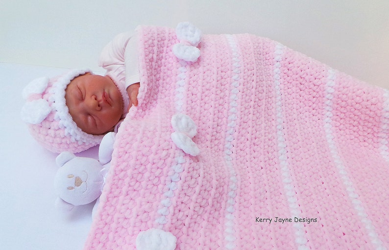 K&ouml;nnte beinhalten: Eine gestrickte Babydecke in rosa und wei&szlig; mit wei&szlig;en geh&auml;kelten Blumen. Die Decke ist &uuml;ber einem Baby gefaltet, das eine rosa und wei&szlig;e geh&auml;kelte M&uuml;tze tr&auml;gt.