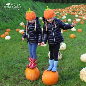 May include: Two children wearing orange and green pumpkin hats stand on top of pumpkins in a field of pumpkins. The children are wearing black puffer jackets, jeans, and rain boots.