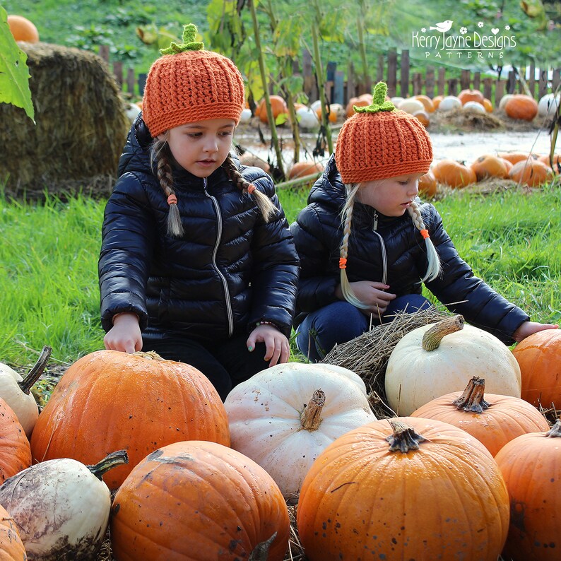 May include: Two children wearing orange pumpkin hats with green stems are sitting in a field of pumpkins. The pumpkins are various sizes and colors, including orange and white.