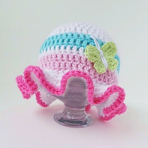 Puede incluir: Un sombrero de sol de ganchillo con una corona a rayas rosa, blanca y azul y un borde de volantes rosa. Una mariposa de ganchillo verde est&aacute; unida al sombrero.