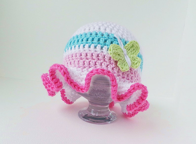 Puede incluir: Un sombrero de sol de ganchillo con una corona a rayas rosa, blanca y azul y un borde de volantes rosa. Una mariposa de ganchillo verde est&aacute; unida al sombrero.