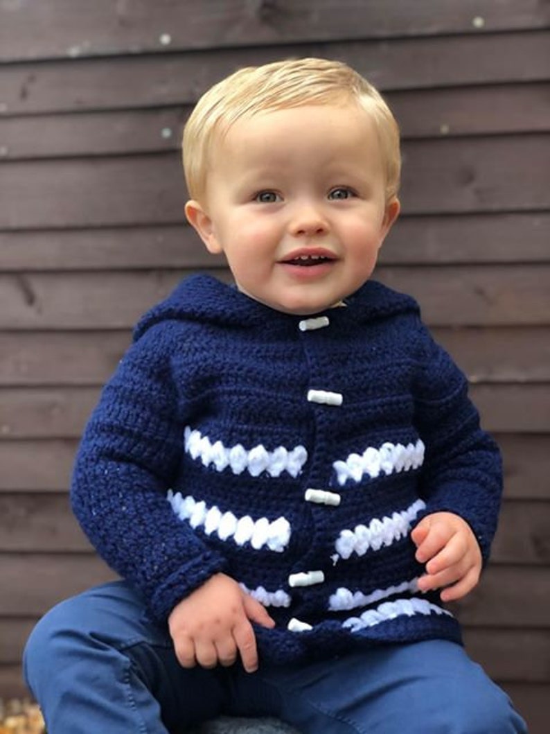 K&ouml;nnte beinhalten: Ein Kleinkind tr&auml;gt eine blaue Strickjacke mit wei&szlig;en Streifen und wei&szlig;en Kn&ouml;pfen. Die Jacke hat eine Kapuze.
