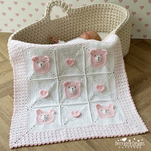 Puede incluir: Una manta de bebé de crochet blanca y rosa con un borde de corazones de crochet rosa y un patrón de osos rosas con caras blancas y corazones rosas. La manta está sobre una cesta de mimbre.