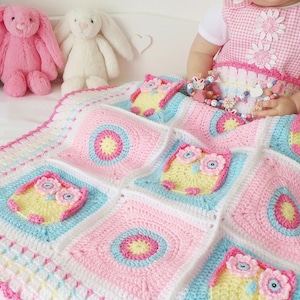 May include: A pink, blue, and white crocheted baby blanket with a border of pink and white crocheted flowers. The blanket features a repeating pattern of owls with yellow bodies, pink flowers, and blue eyes. The blanket is on a white bed with three stuffed bunnies.