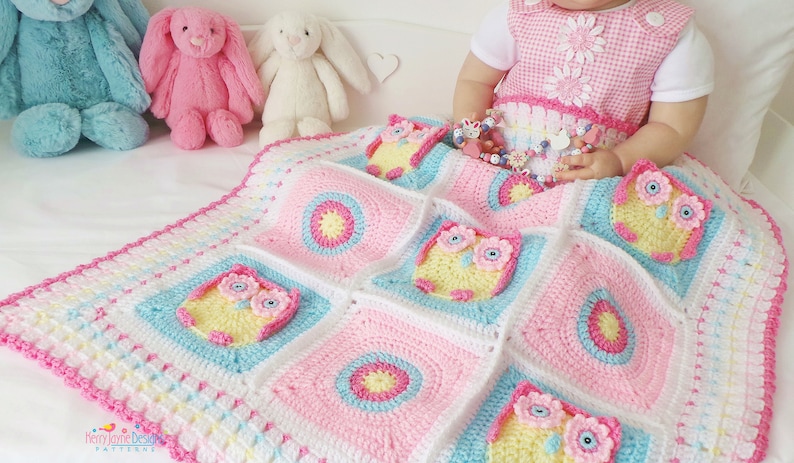May include: A pink, blue, and white crocheted baby blanket with a border of pink and white crocheted flowers. The blanket features a repeating pattern of owls with yellow bodies, pink flowers, and blue eyes. The blanket is on a white bed with three stuffed bunnies.