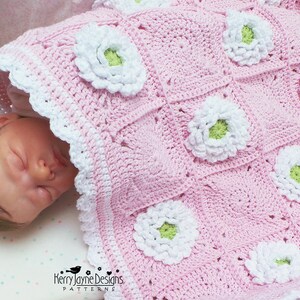 May include: A pink crocheted baby blanket with white and green flowers. The blanket is made with a granny square pattern and has a white crocheted border.