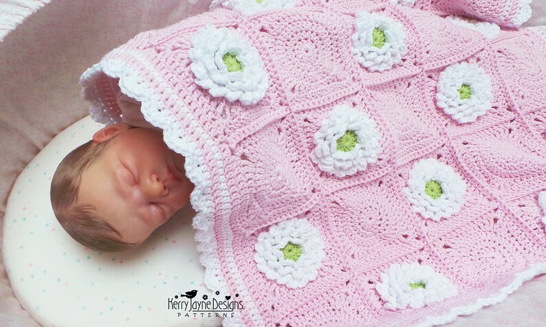 May include: A pink crocheted baby blanket with white and green flowers. The blanket is made with a granny square pattern and has a white crocheted border.