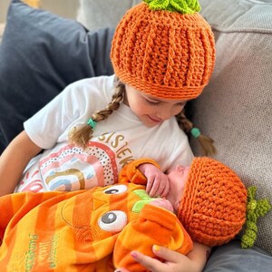 May include: Two babies wearing orange and green crocheted pumpkin hats. The older baby is holding the younger baby. The older baby is wearing a white shirt with the word "Sister" on it. The younger baby is wearing an orange and green pumpkin onesie.