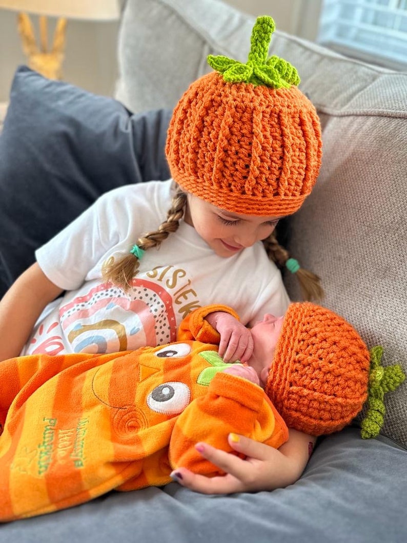 May include: Two babies wearing orange and green crocheted pumpkin hats. The older baby is holding the younger baby. The older baby is wearing a white shirt with the word "Sister" on it. The younger baby is wearing an orange and green pumpkin onesie.