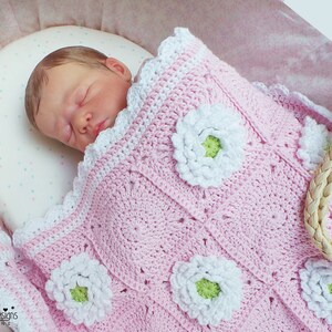 May include: A pink crocheted baby blanket with white daisy flowers. The blanket has a scalloped edge and is made with a soft yarn.