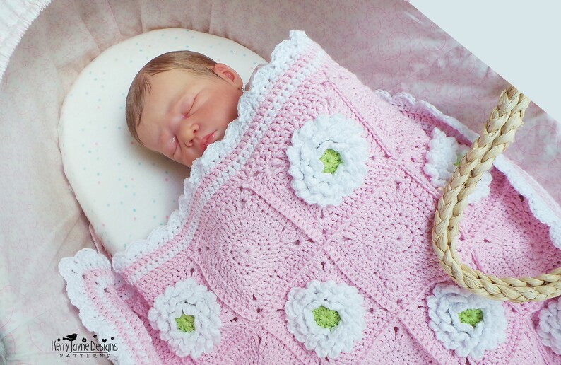 May include: A pink crocheted baby blanket with white daisy flowers. The blanket has a scalloped edge and is made with a soft yarn.